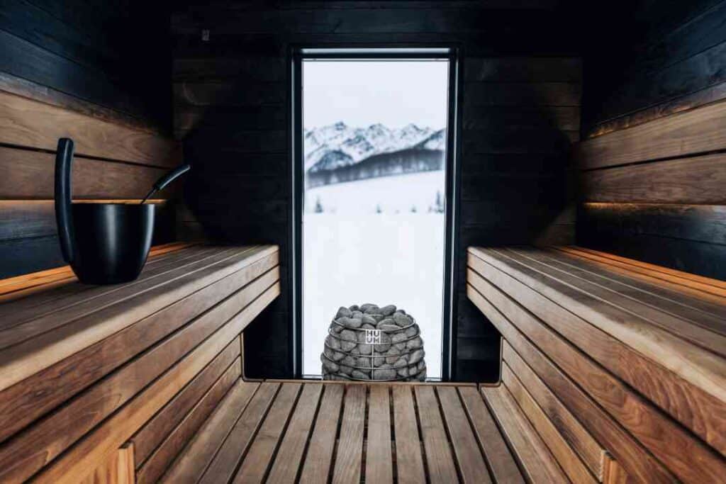 Photo from inside a sauna, with wooden benches, a rock filled oven, and a window to the winter snow outside the sauna