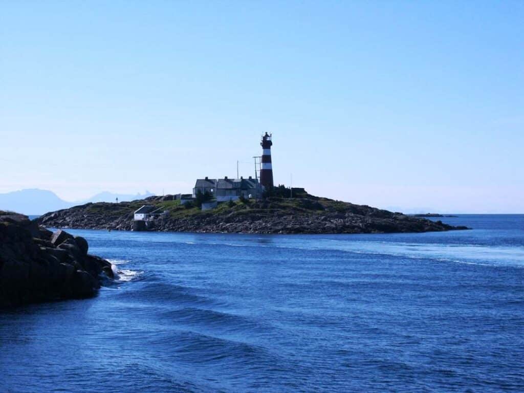 A red and white lighthouse alone on a small island surrounded by deep blue sea under a pale blue sky in Lofoten Norway, with mountains vaguely visible in the distance across the water