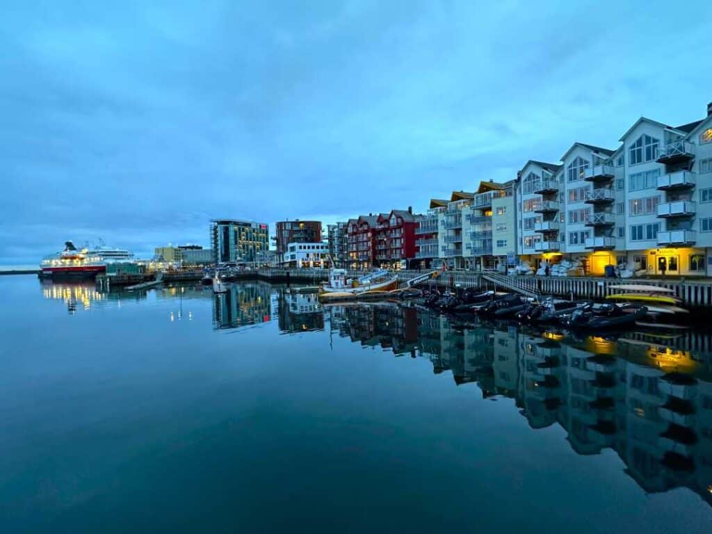 The town dock in Svolvær at dusk, with lots of traditional houses in different colors along the dock, boats on the blank harbor water, and warm lighting from buildings and boats under a grey sky.