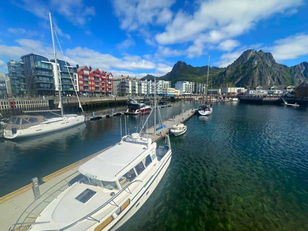 A couple of white boats by the dock in a harbor with greenish clear water, colorful buildings along the dock and majestic mountains in the background under a blue sky, in summer in Svolvær Lofoten Norway.