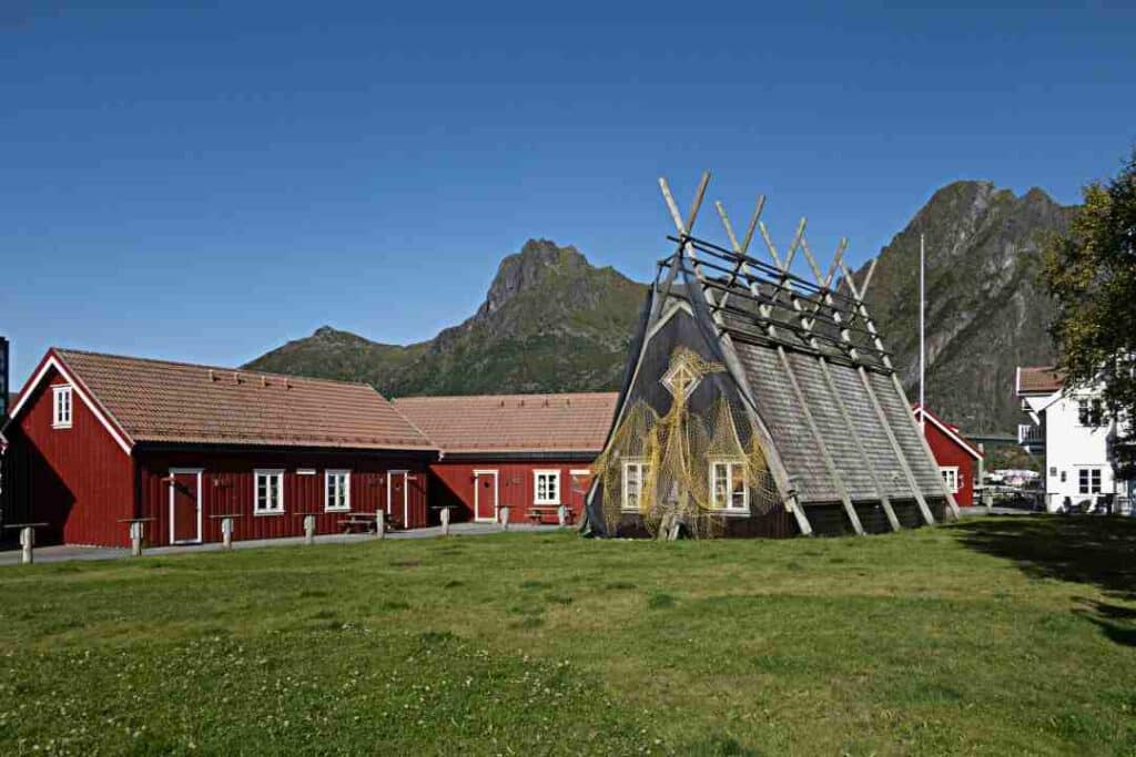 Red fishermans cabin and a traditional barn with stockfish racks on a green field under majestic mountains on a bright sunny day in Svolvær Lofoten, Norway.