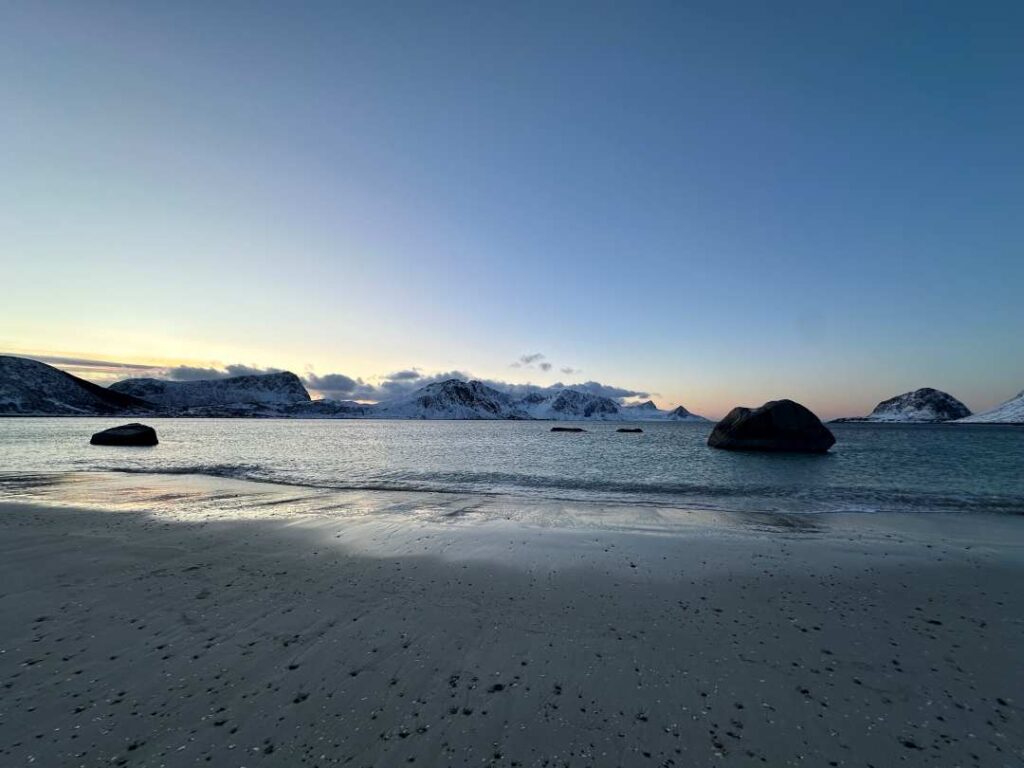 A white ssndy winter breach in front of a dark but silvery ocean, snow covered mountains in the distance across the water under a pale blue winter sky